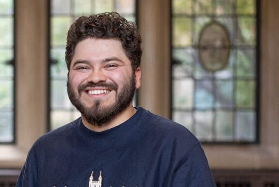 A bearded person smiles in front of stained glass windows in a Michigan Law sweatshirt.